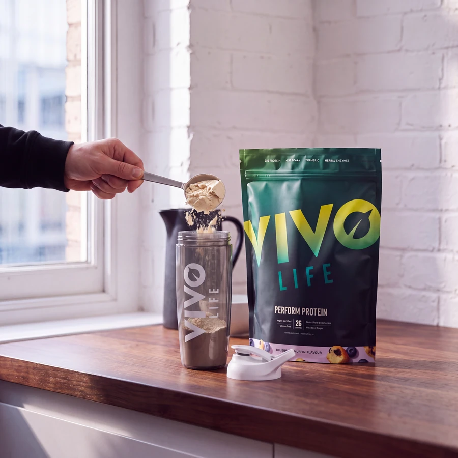 Krok 2: wsypywanie odmierzonej porcji PERFORM jagodowy muffin łyżką stołową do shakera Vivo Life, obok opakowanie 936 g